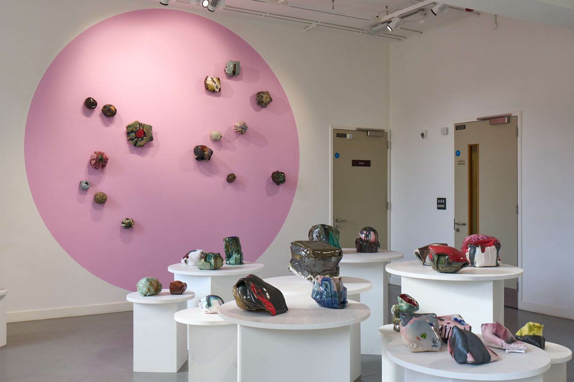 A gallery space with large pink circle painted on the wall. On this wall and plinths below are lots of brightly coloured and irregularly shaped ceramic sculptures.